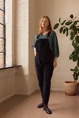 Woman wearing black overalls standing in a room with a plant and window.