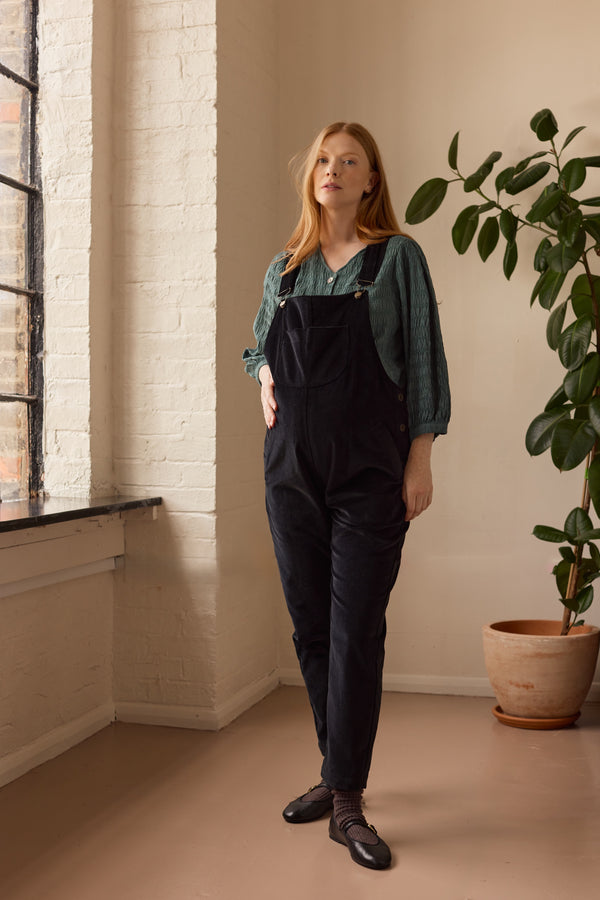 Woman wearing black overalls standing in a room with a plant and window.