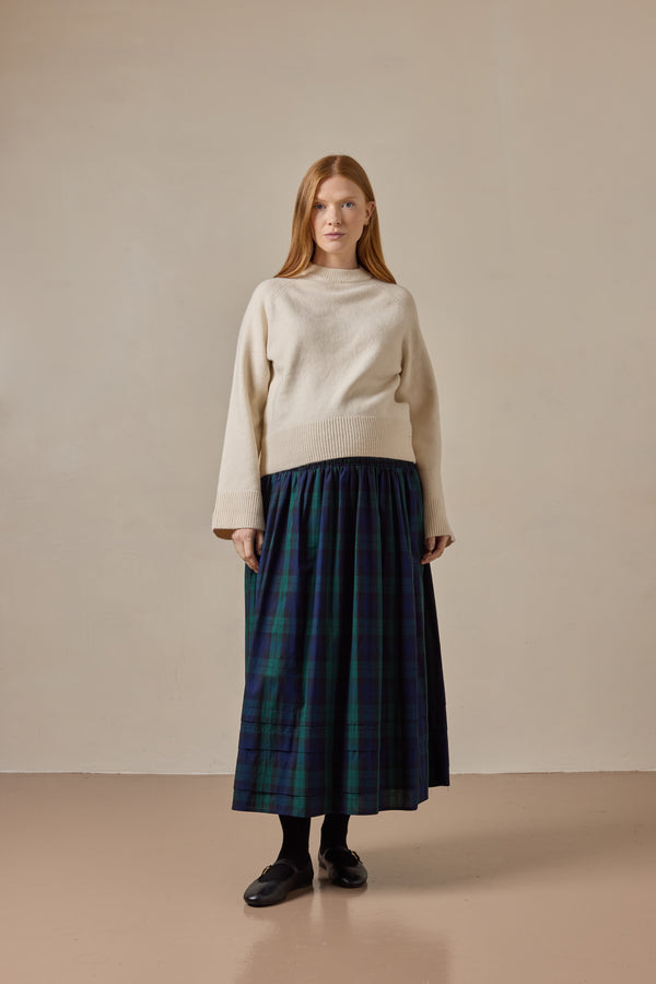 Woman wearing a cream sweater and green and blue tartan skirt against a beige background
