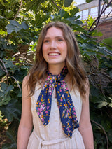 A smiling young woman with long light brown hair stands outdoors in front of leafy green plants, wearing a cream sleeveless dress and a navy neck scarf with a colorful floral pattern.