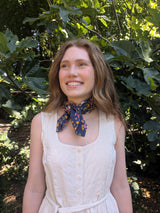 A woman with light brown hair smiling outdoors in front of green foliage, wearing a cream-colored sleeveless dress and a navy blue neck scarf with a colorful floral pattern.