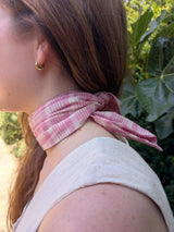 A close-up side profile of a woman wearing a pink and white plaid neck scarf tied behind her neck, with green foliage in the background.